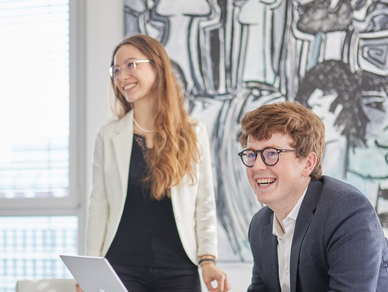 Ein Mann sitzt lächelnd am Laptop, während eine Frau neben ihm steht und ebenfalls lächelt. Beide befinden sich in einem modernen Büro mit kunstvollen Wandgemälden.