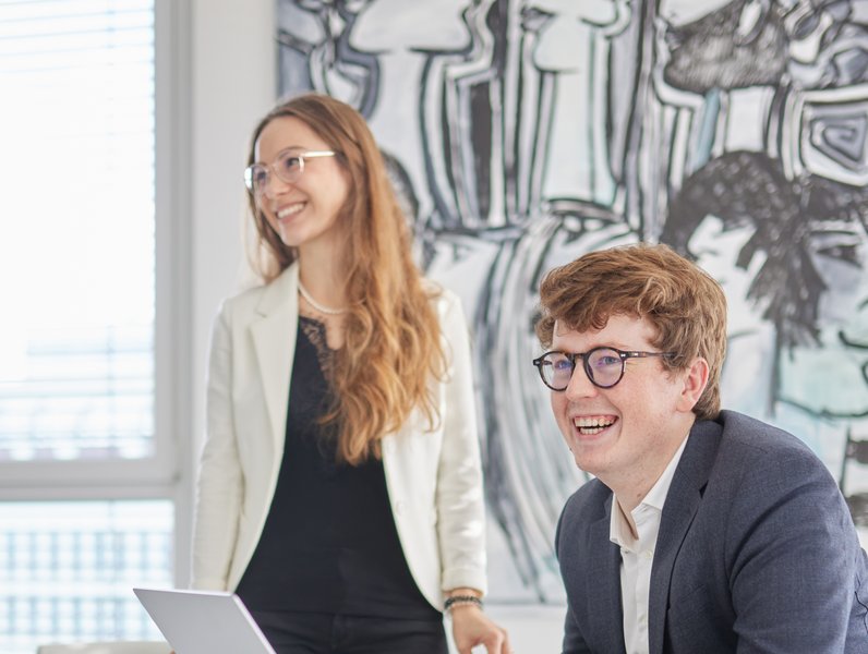 Ein Mann sitzt lächelnd am Laptop, während eine Frau neben ihm steht und ebenfalls lächelt. Beide befinden sich in einem modernen Büro mit kunstvollen Wandgemälden.