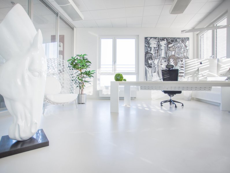 Eine weiße Pferdeskulptur steht in einem modernen, hellen Büro mit einem Schreibtisch und Bürostuhl vor einem großen Fenster, einem Kunstwerk an der Wand und einer Zimmerpflanze.