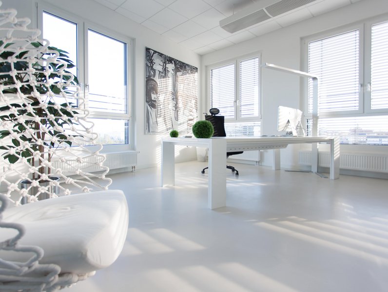 Ein moderner, weißer Schreibtisch steht in einem hellen Büro mit großen Fenstern und Jalousien. Eine schwarze Bürostuhl, grüne Pflanzenkugel und Kunst an der Wand ergänzen die minimalistische Einrichtung.