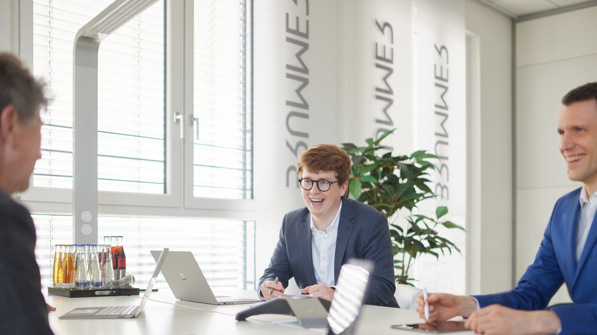 Eine Gruppe von Männern lacht und arbeitet an Laptops in einem modernen Konferenzraum mit Pflanzen, Getränken und dem Text "3NWWE5" auf Schirmen an den Fenstern.