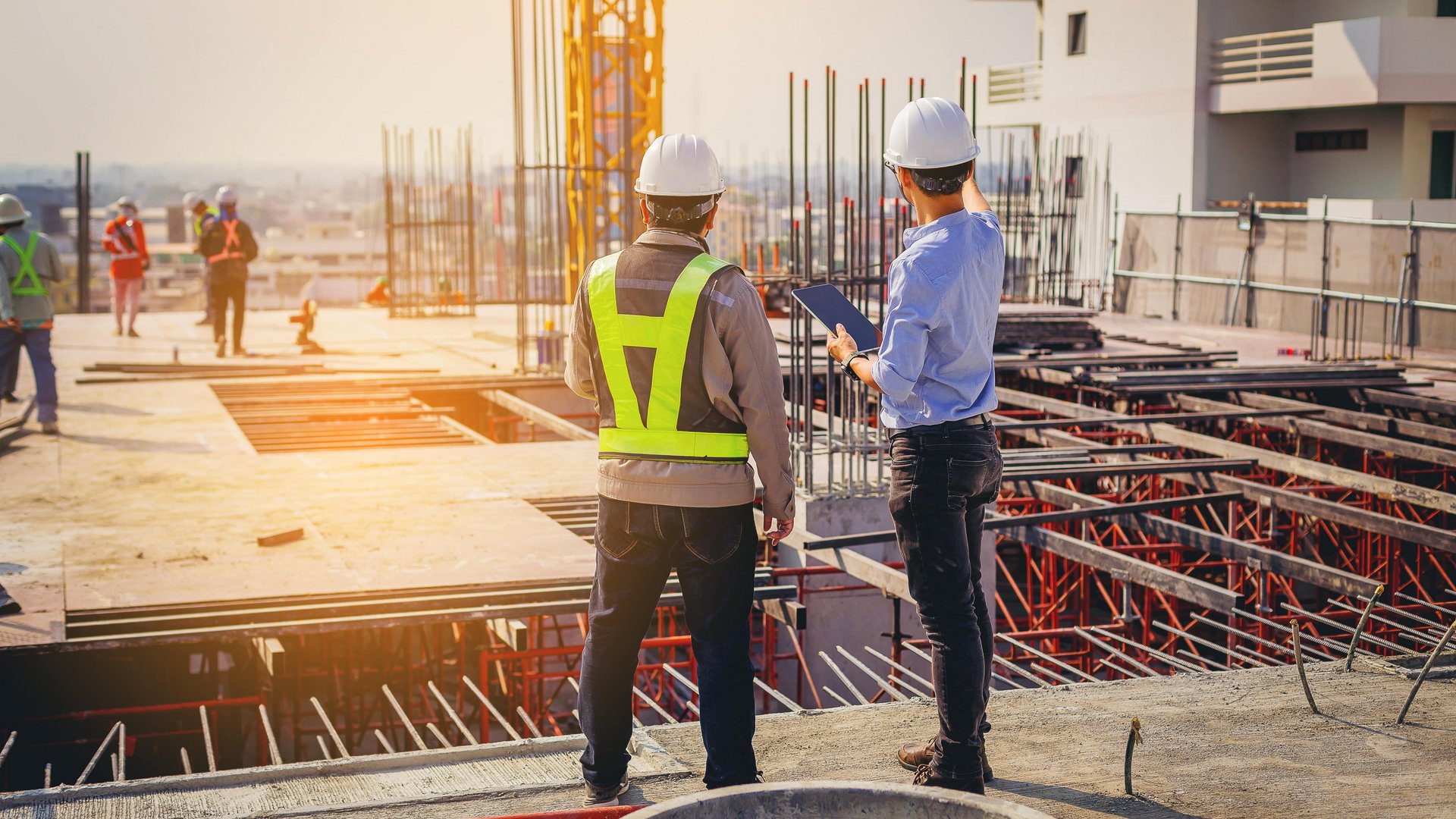 Zwei Bauarbeiter tragen Schutzhelme und Westen, stehen nebeneinander und untersuchen Pläne auf einer Baustelle. Im Hintergrund sind weitere Arbeiter und Baugerüste zu sehen, bei Tageslicht.
