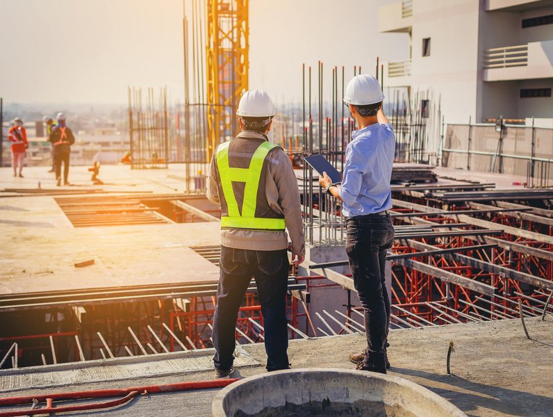 Zwei Bauarbeiter tragen Schutzhelme und Westen, stehen nebeneinander und untersuchen Pläne auf einer Baustelle. Im Hintergrund sind weitere Arbeiter und Baugerüste zu sehen, bei Tageslicht.
