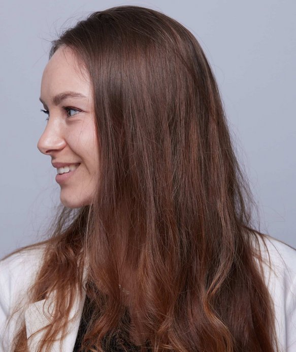 Eine Frau mit langen, braunen Haaren lächelt und schaut nach links, vor einem schlichten grauen Hintergrund. Sie trägt ein weißes Kleidungsstück.