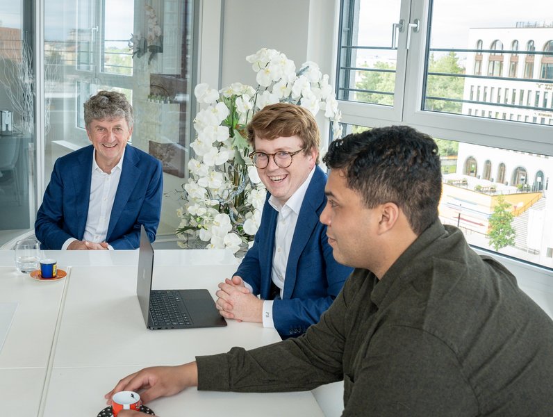 Drei Männer sitzen lächelnd an einem Besprechungstisch mit einem Laptop. Im Hintergrund sind große Fenster und Orchideenblumen zu sehen, das Tageslicht hellt den Raum auf.