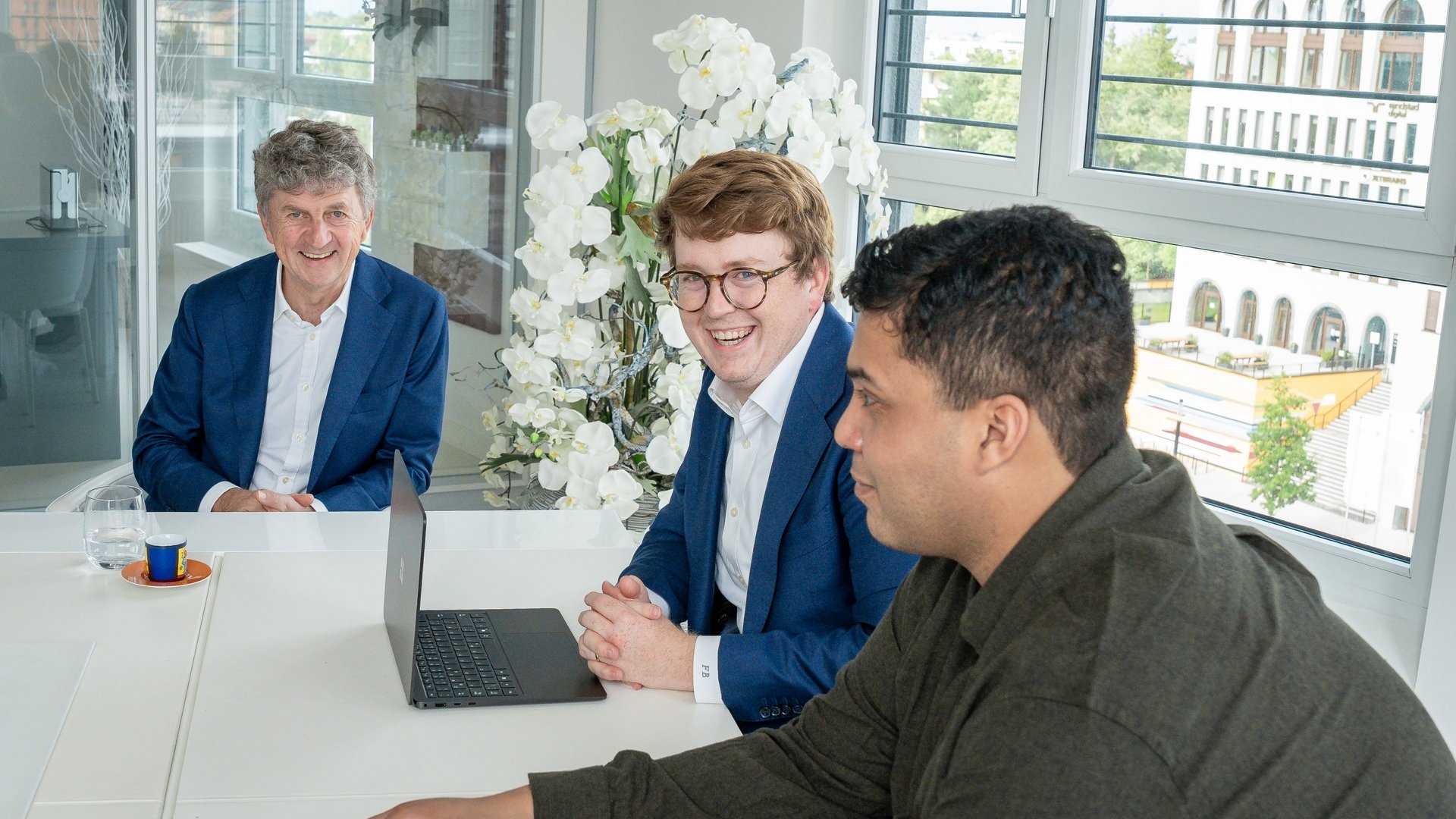 Drei Männer sitzen lächelnd an einem Besprechungstisch mit einem Laptop. Im Hintergrund sind große Fenster und Orchideenblumen zu sehen, das Tageslicht hellt den Raum auf.