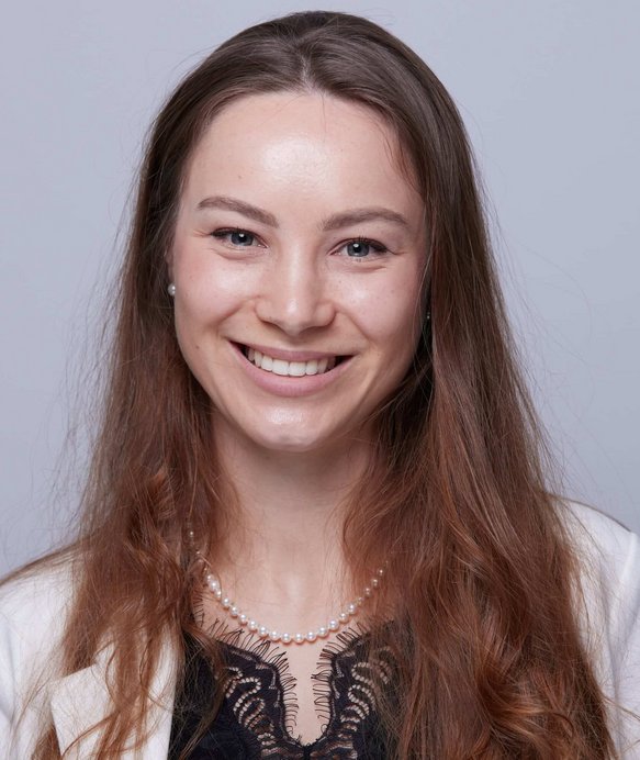 Eine Frau lächelt in die Kamera, trägt eine Perlenkette und eine weiße Jacke, im Hintergrund ein neutraler grauer Hintergrund.