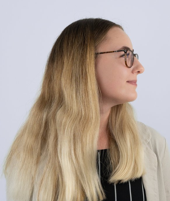 Eine Frau mit langen blonden Haaren und Brille blickt nach oben. Sie trägt einen schwarzen, gestreiften Pullover und befindet sich vor einem neutralen, hellen Hintergrund.