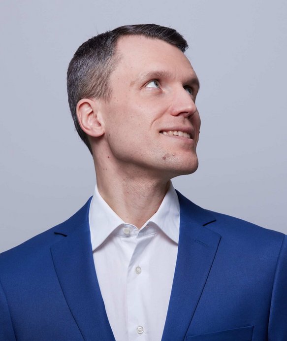 Ein Mann in einem blauen Anzug schaut lächelnd nach oben, vor einem neutralen grauen Hintergrund.