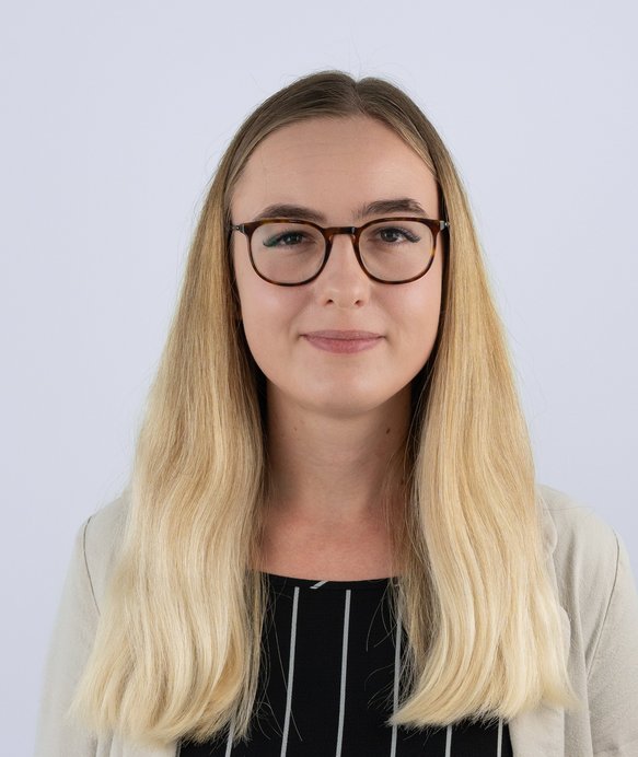 Eine Frau mit blonden Haaren und Brille blickt in die Kamera. Sie trägt ein gestreiftes Oberteil und eine helle Jacke, vor einem neutralen Hintergrund stehend.