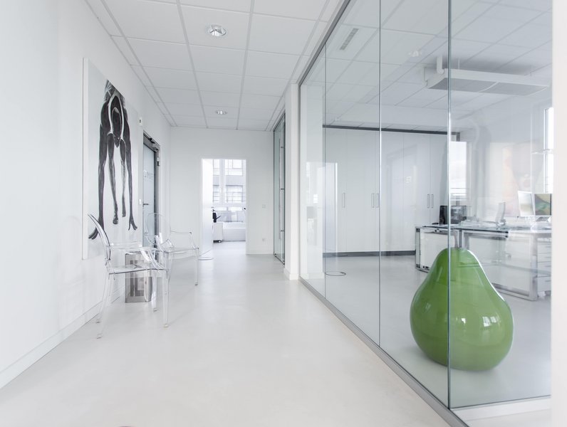 Ein moderner Flur mit gläserner Trennwand, daneben ein grüner birnenförmiger Sitz. Transparentstühle stehen entlang der Wand, geschmückt mit einem schwarz-weißen Gemälde. Helle, minimalistische Büroumgebung.