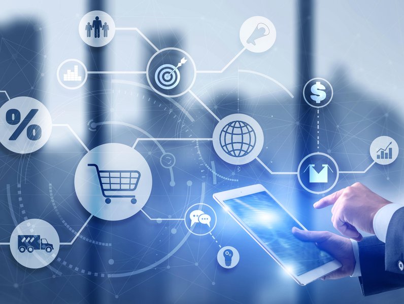 Ein Tablet wird von einer Hand bedient und zeigt digitale Symbole wie Einkaufswagen, Weltkugel und Zielscheibe vor verschwommenem Hintergrund. Symbole stehen für Kommunikation, Finanzen und Globalisierung.