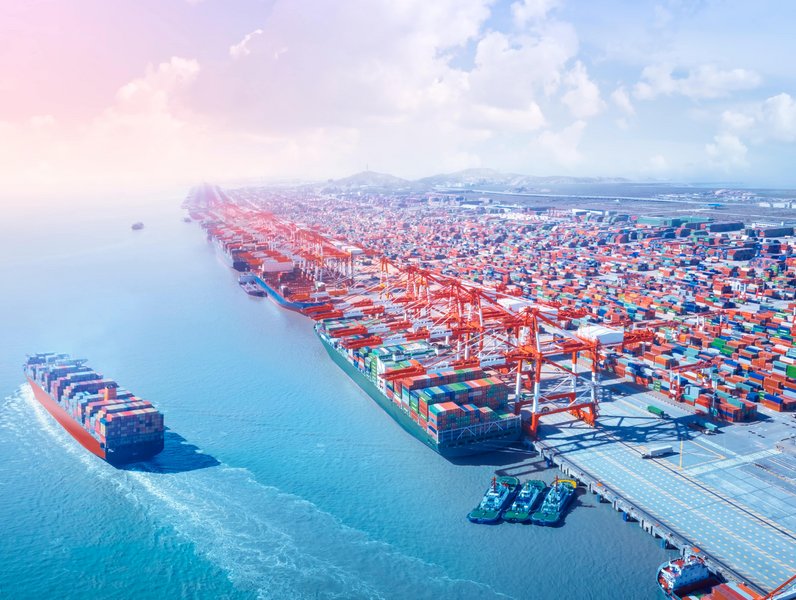 Ein Containerschiff verlässt einen belebten Hafen, während unzählige Container aufgestapelt sind. Kräne und Schiffe dominieren die Szene unter einem klaren, wolkigen Himmel entlang des Wassers.