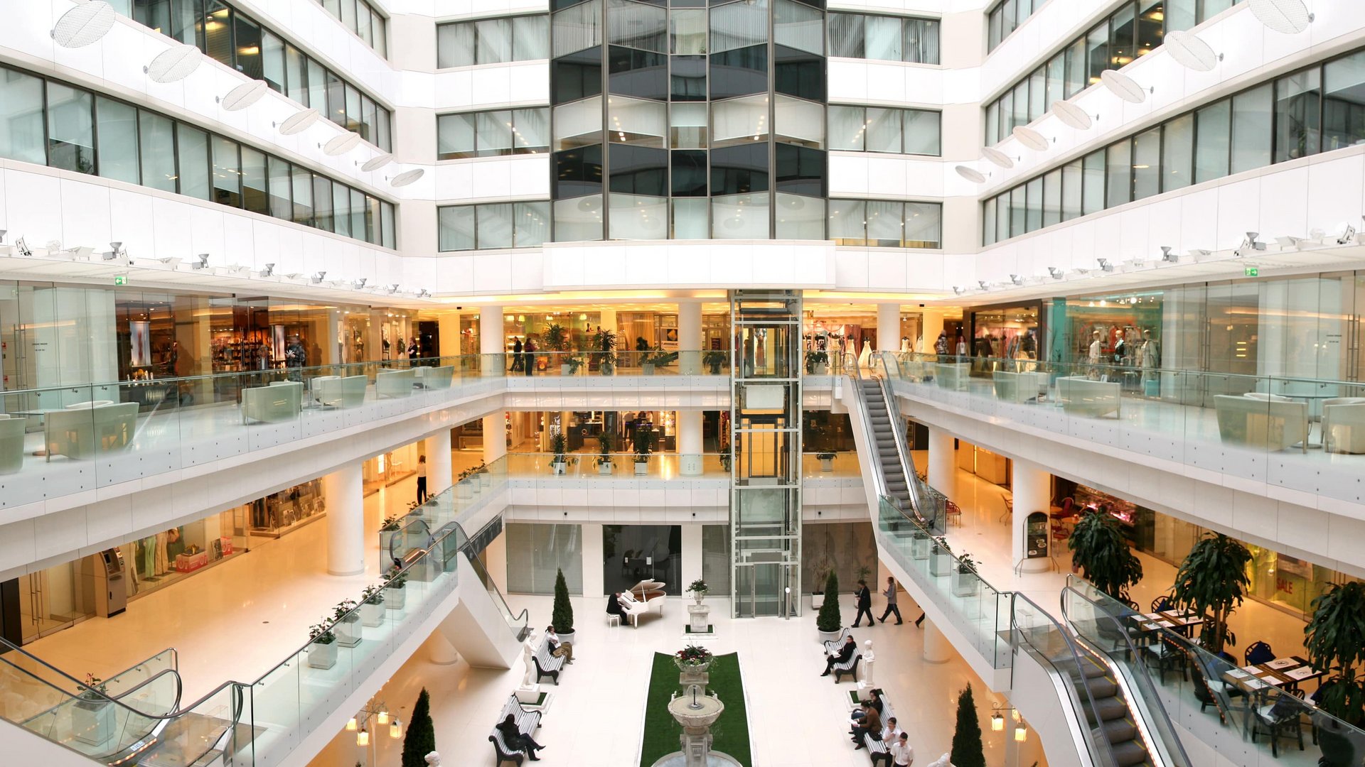 Ein modernes Einkaufszentrum mit mehreren Stockwerken, gläsernen Geländern und Rolltreppen. Besucher schlendern vorbei an Geschäften, während Sitzbereiche im Erdgeschoss zur Entspannung einladen.