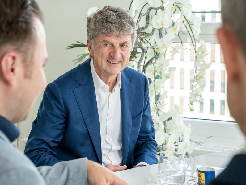 Ein Mann im blauen Anzug lächelt, während er an einem Tisch mit zwei anderen Personen sitzt. Im Hintergrund sind weiße Orchideen vor einem Fenster sichtbar.