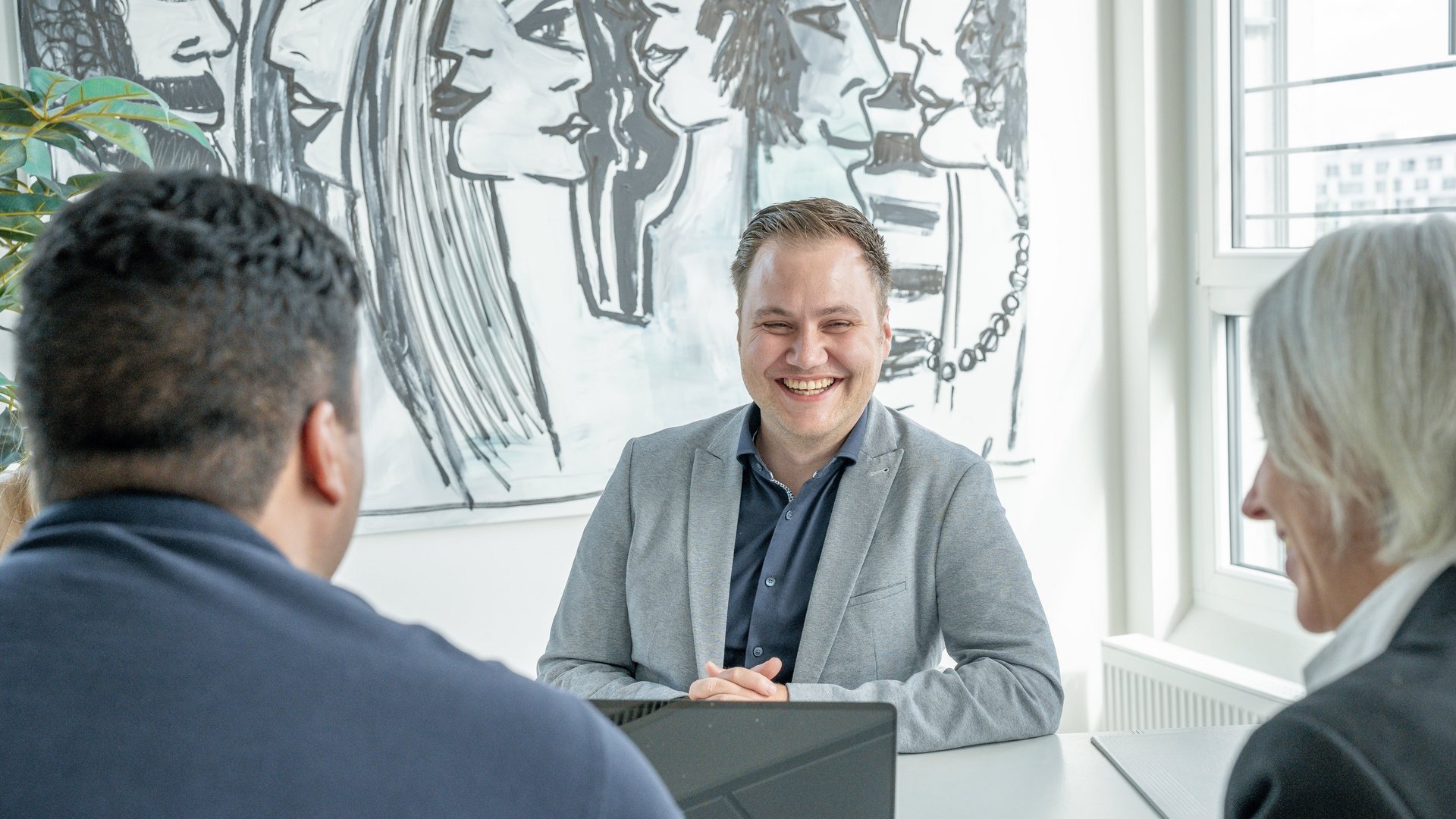 Ein Mann in einem grauen Anzug lächelt, während er an einem Tisch sitzt. Zwei Personen ihm gegenüber. Im Hintergrund befindet sich ein schwarz-weißes Wandbild.