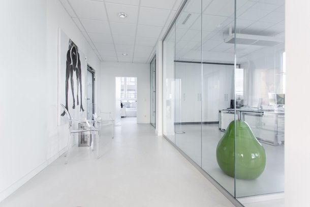 Ein moderner Flur mit gläserner Trennwand, daneben ein grüner birnenförmiger Sitz. Transparentstühle stehen entlang der Wand, geschmückt mit einem schwarz-weißen Gemälde. Helle, minimalistische Büroumgebung.