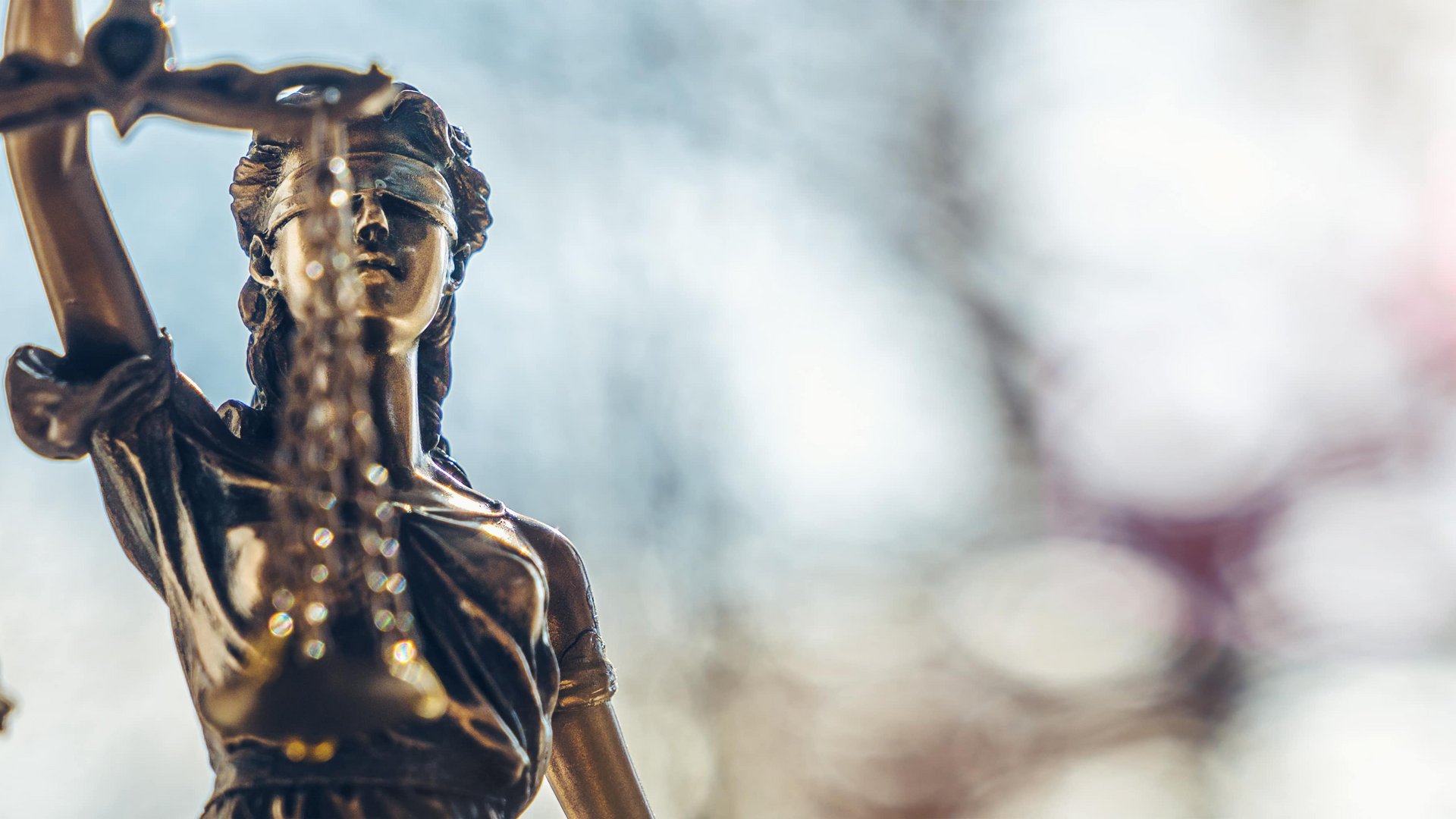 Eine Justitia-Statue hält eine Waage hoch, symbolisch für Gerechtigkeit. Der Hintergrund ist unscharf, mit diffusen Lichtern und Farben, die eine ruhige Atmosphäre schaffen.