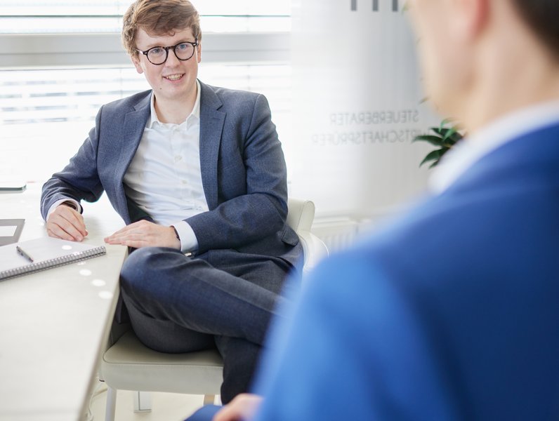 Eine Person sitzt lächelnd in einem Anzug am Schreibtisch, hält ein Notizbuch, in einem hellen Büro. Im Hintergrund sind Pflanzen und verschwommener Schriftzug sichtbar.