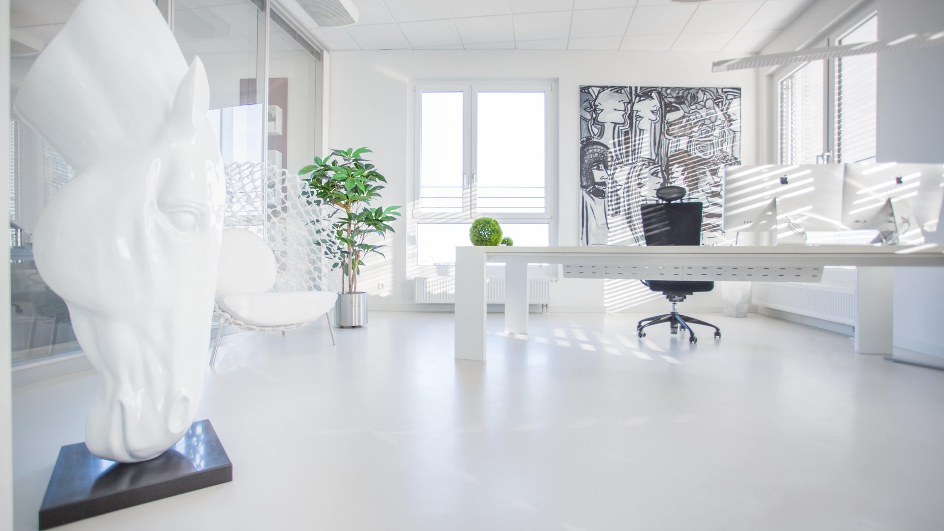 Eine weiße Pferdeskulptur steht in einem modernen, hellen Büro mit einem Schreibtisch und Bürostuhl vor einem großen Fenster, einem Kunstwerk an der Wand und einer Zimmerpflanze.