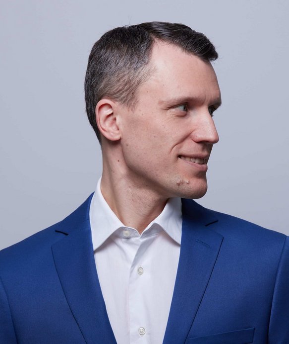 Ein Mann in einem blauen Anzug und weißem Hemd schaut zur Seite, lächelnd. Der Hintergrund ist einfarbig grau, was den Fokus auf seine Gesichtsausdrücke und Kleidung lenkt.