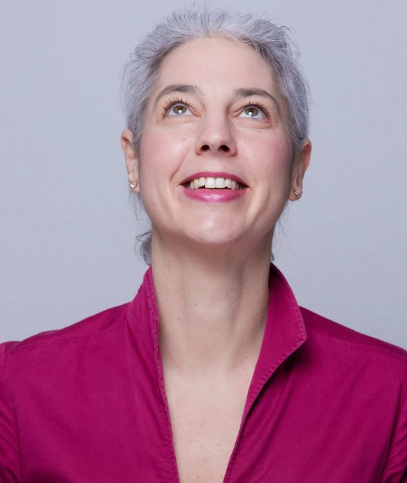 Eine Frau mit grauen Haaren, lächelt und blickt nach oben, trägt eine pinke Bluse, vor einem neutralen, hellgrauen Hintergrund.