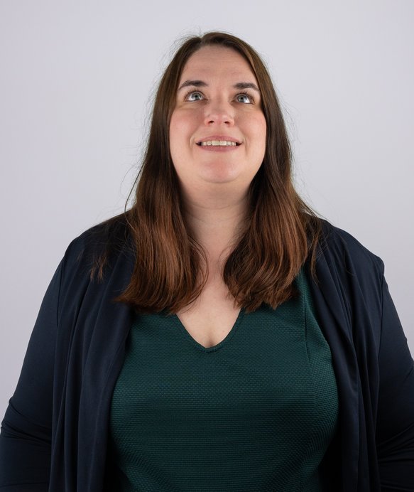 Frau mit braunen Haaren sucht lächelnd nach oben. Sie trägt ein grünes Oberteil und einen dunklen Blazer. Der Hintergrund ist schlicht und weiß.