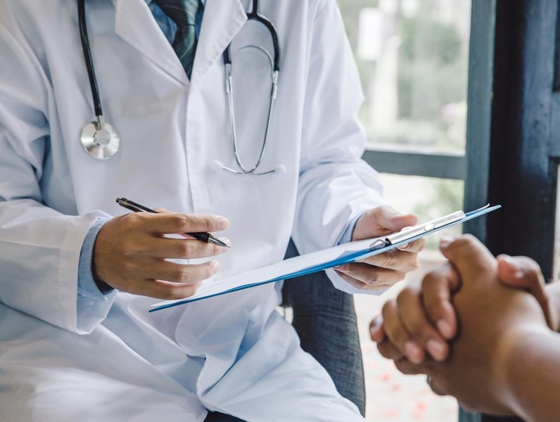 Ein Arzt mit Stethoskop schreibt auf ein Klemmbrett. Gegenüber sitzt eine Person mit gefalteten Händen. Der Raum ist hell mit Fenstern im Hintergrund.