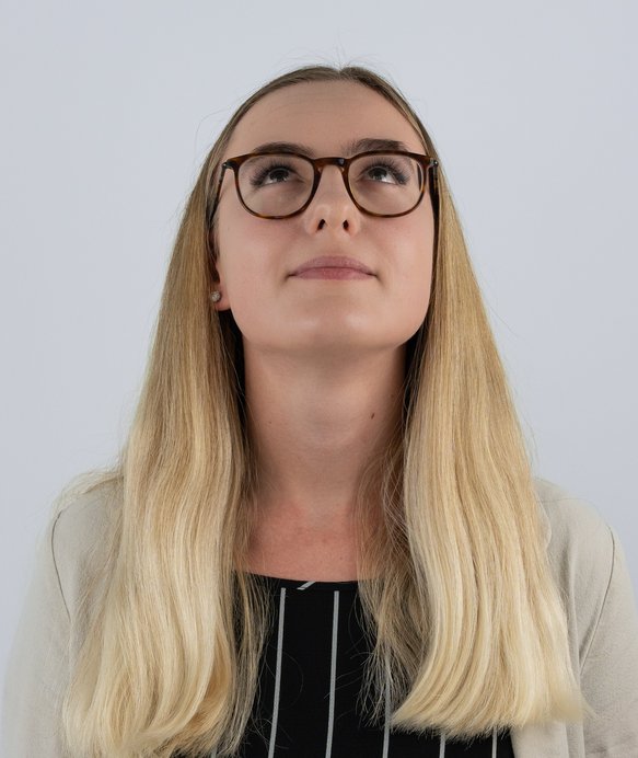 Eine Frau mit langen blonden Haaren und Brille schaut nach oben. Sie trägt ein gestreiftes Oberteil unter einem beigen Cardigan und steht vor einem neutralen, hellgrauen Hintergrund.