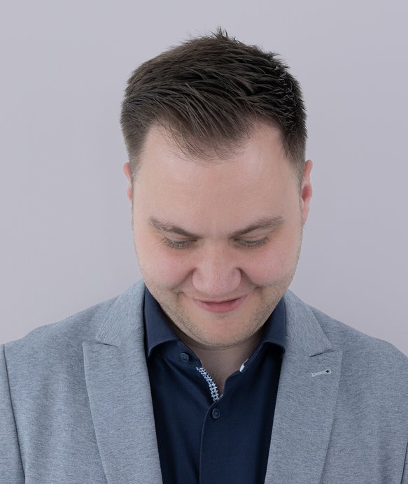 Ein Mann in einem grauen Anzug schaut nach unten und lächelt leicht. Der Hintergrund ist einheitlich hellgrau, was die konzentrierte und ruhige Stimmung verstärkt.