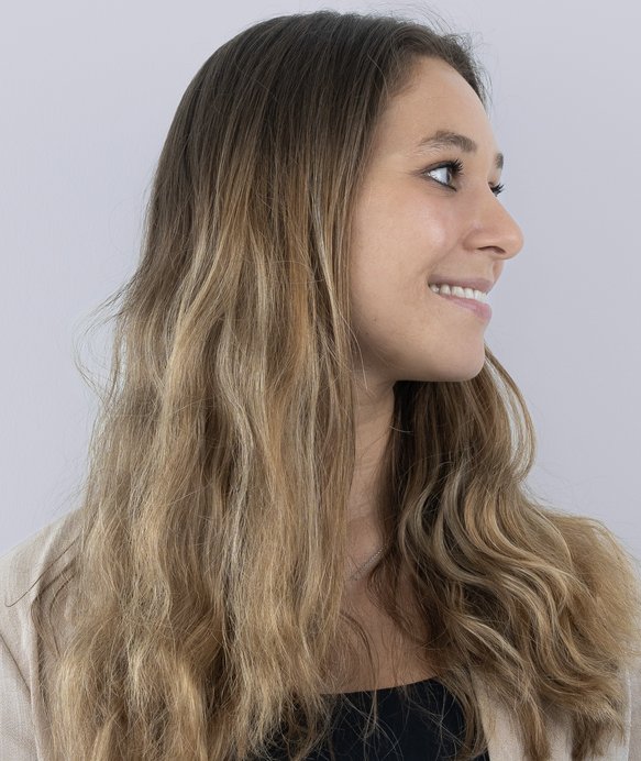 Eine Frau mit langen, welligen Haaren schaut lächelnd zur Seite. Sie trägt eine helle Kleidung und steht vor einem neutralen, grauen Hintergrund.