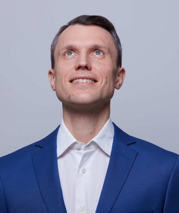 Ein Mann in einem blauen Anzug schaut lächelnd nach oben. Der Hintergrund ist neutral hellgrau.