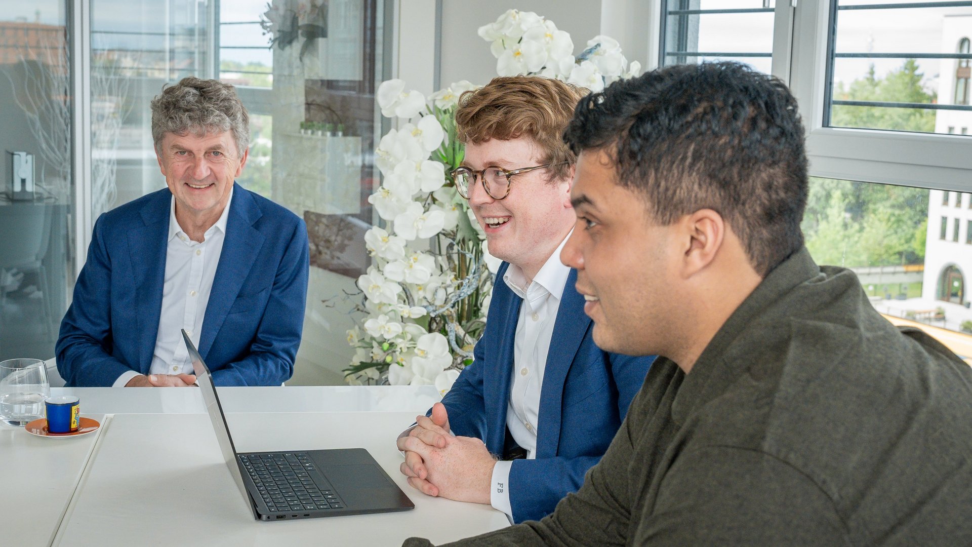 Drei Männer sitzen an einem Tisch, lächeln und sprechen. Ein Laptop steht vor ihnen. Im Hintergrund sind große Fenster und weiße Blumen zu sehen, die eine helle, freundliche Atmosphäre schaffen.