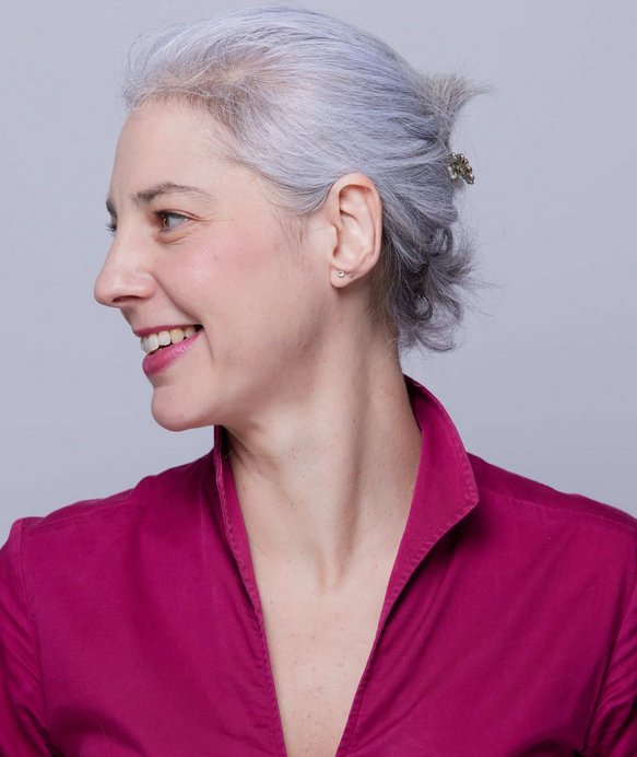 Eine Frau mit grauem Haar lächelt und schaut nach links. Sie trägt eine fuchsiafarbene Bluse. Der Hintergrund ist einfarbig hellgrau.