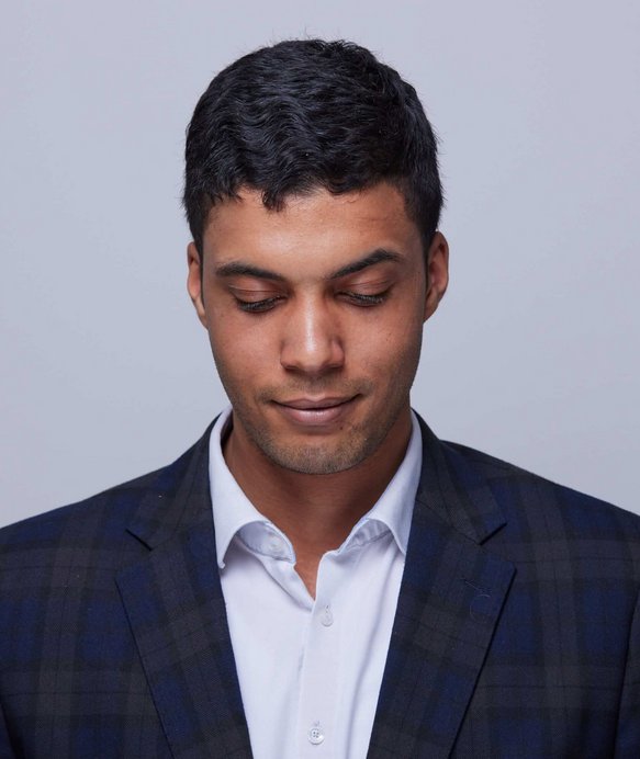 Ein Mann mit dunklen Haaren schaut nach unten. Er trägt ein kariertes Sakko über einem weißen Hemd. Der Hintergrund ist schlicht und grau.